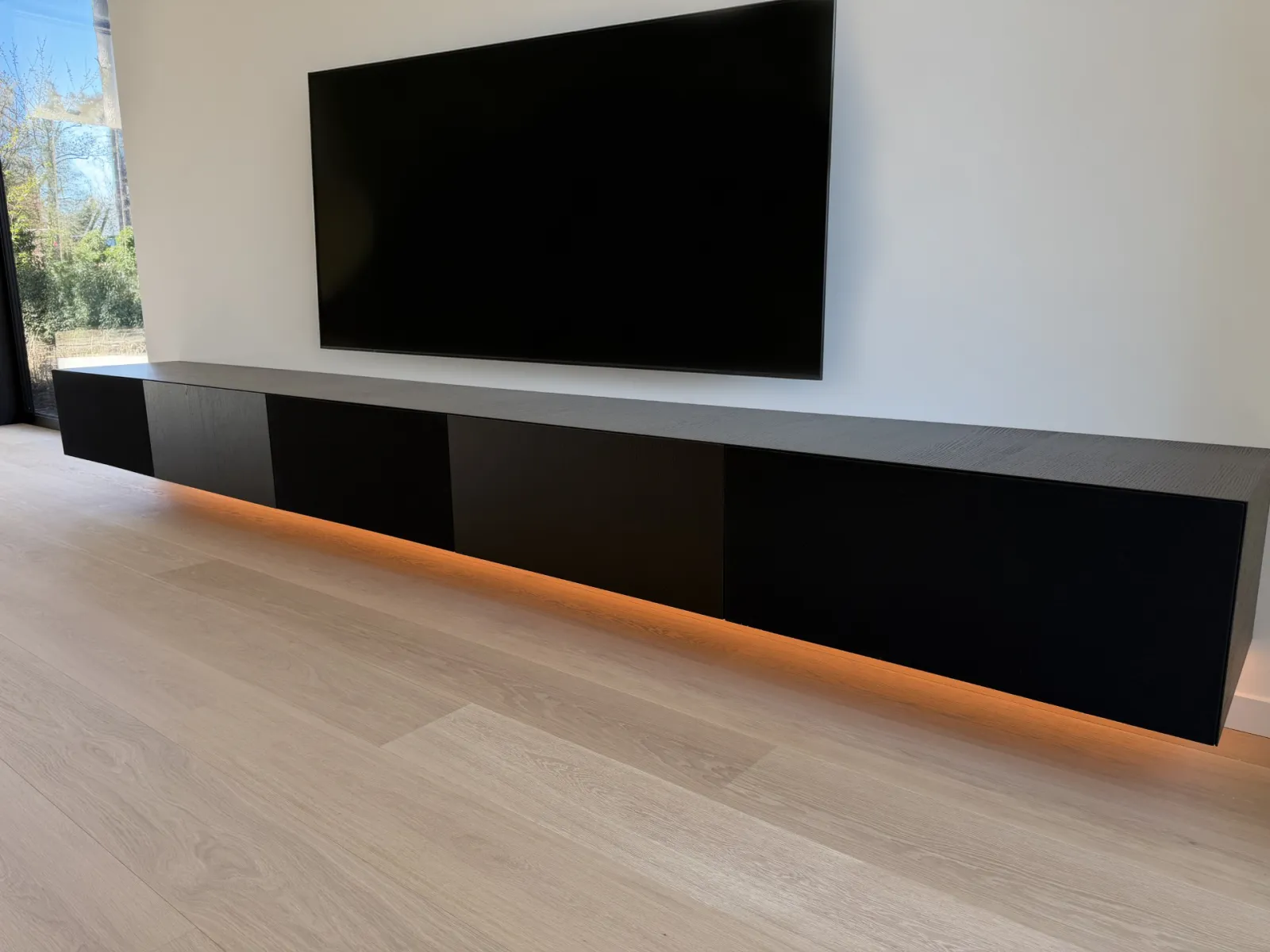 Zwevend Ardeno tv meubel in zwart, met een warme ledlijn onder het meubel en de televisie aan de muur