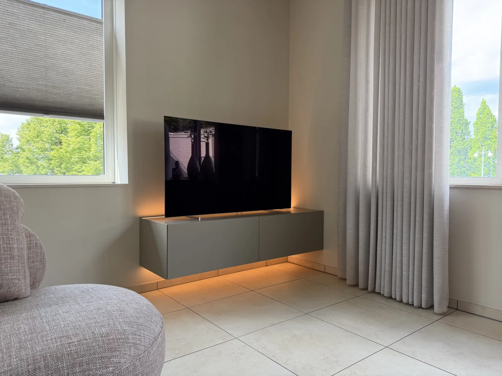 Lichtgrijs JOY! tv meubel met een warme ledlijn eronder, tussen een raam en een gordijn
